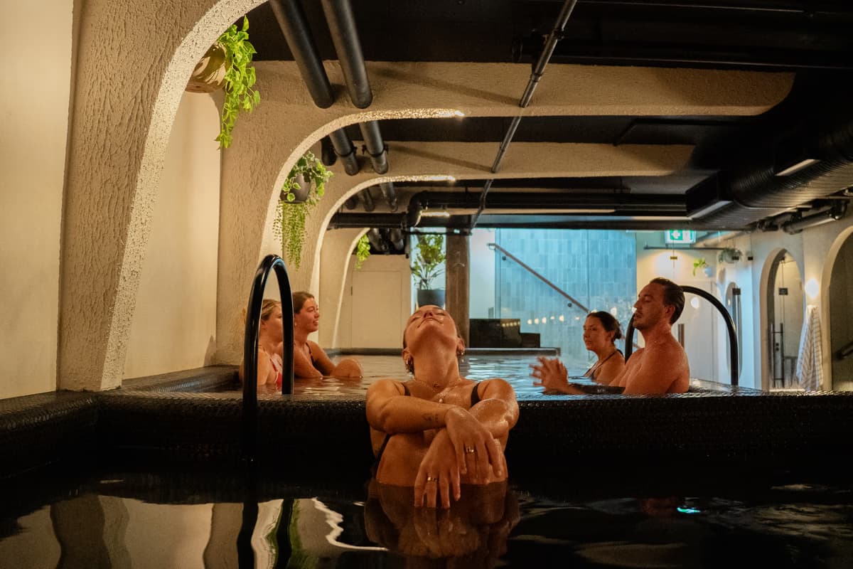 Bathers resting legs raised in a dim cold plunge, the Soak stage of the Navia ritual